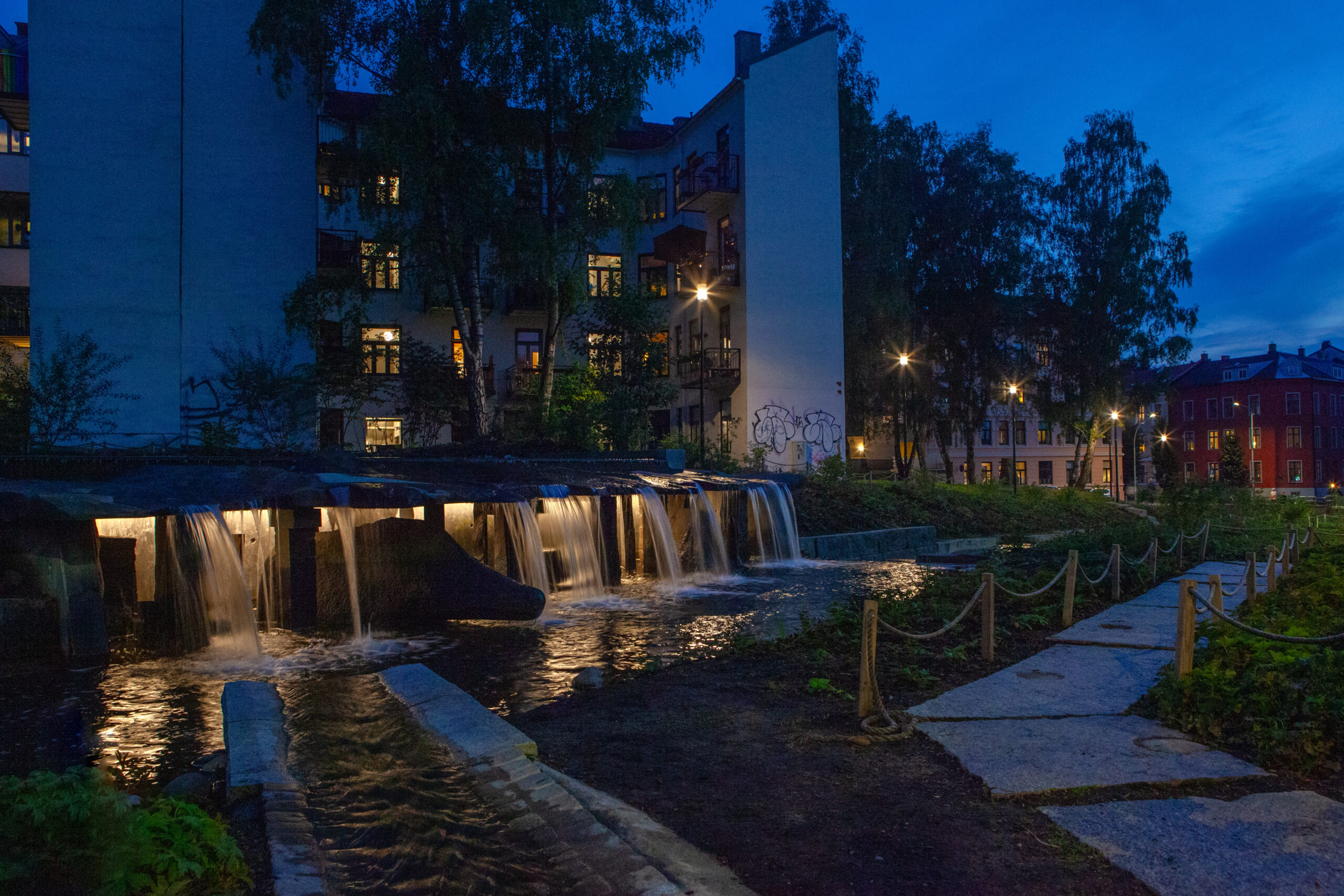 Night view of Klosterenga Park in Oslo, featuring illuminated water cascades flowing through stone structures. The park's serene lighting highlights the natural elements, with the soft glow from beneath the waterfalls complementing the urban surroundings and creating a tranquil ambiance. Trees and pathways are dimly lit in the background, blending art, nature, and lighting design seamlessly.