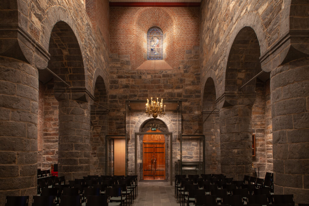 Interior of Gamle Aker Church in Oslo, with warm lighting accentuating the Romanesque stone arches and creating a serene, historic ambiance. The chandelier and hidden lighting enhance the space while maintaining focus on the architecture.