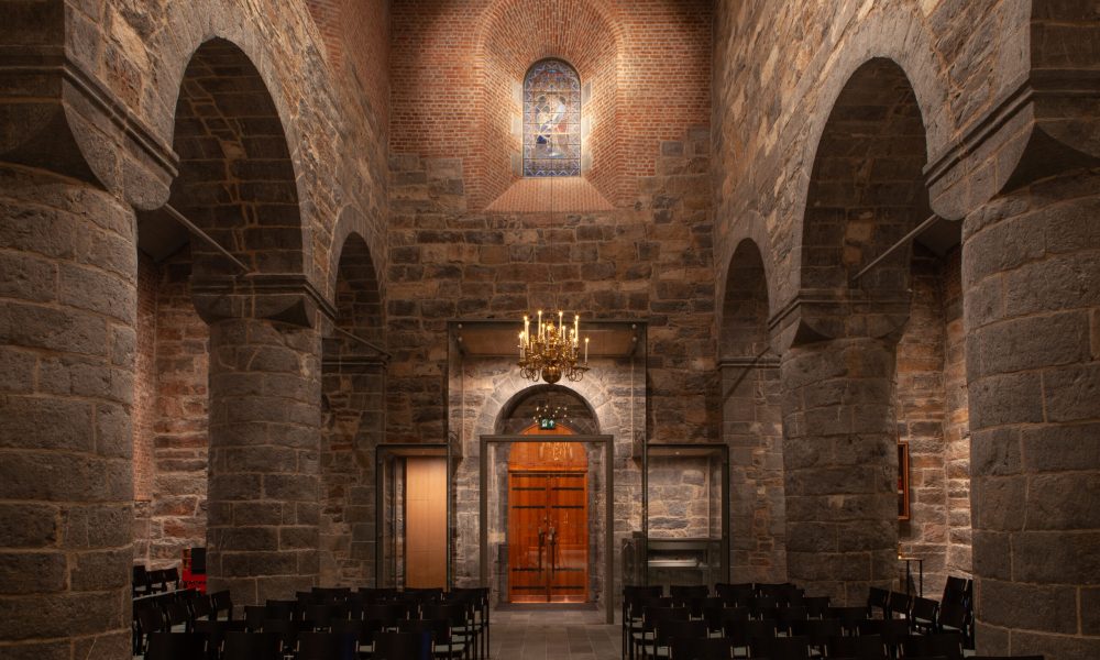 Interior of Gamle Aker Church in Oslo, with warm lighting accentuating the Romanesque stone arches and creating a serene, historic ambiance. The chandelier and hidden lighting enhance the space while maintaining focus on the architecture.